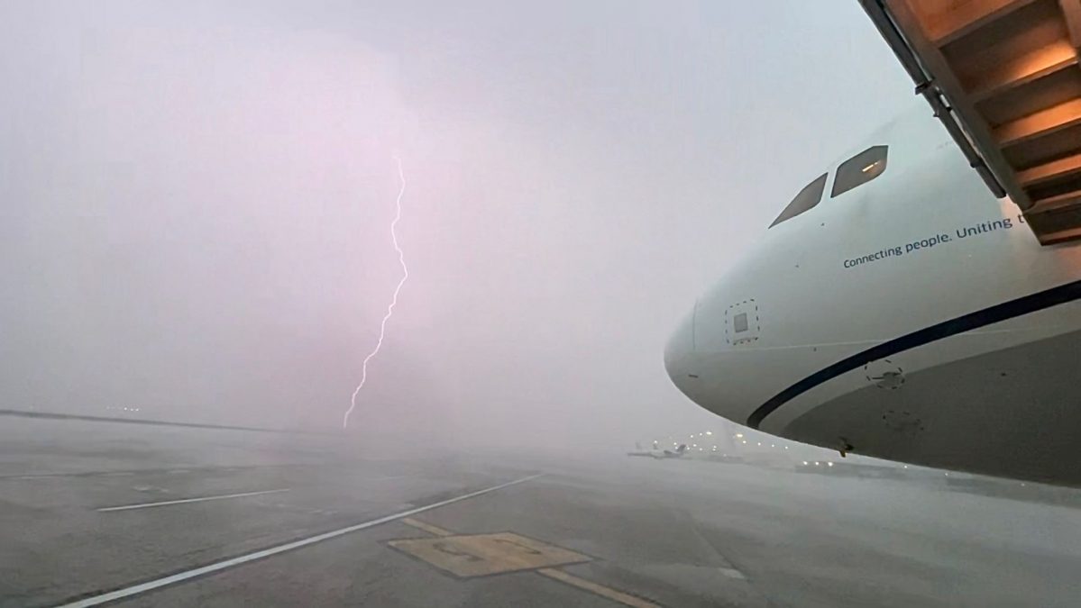 Photograph taken on a stormy day at St. Louis Lambert International Airport, showing a lightning bolt striking in the distance across a foggy, rain-soaked tarmac. The front of a United Airlines aircraft is visible on the right side of the image, with the slogan 'Connecting people. Uniting the world.' partially readable along the fuselage.
