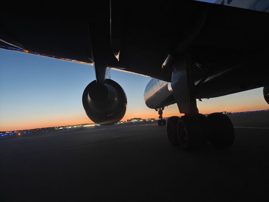 Low-angle photograph taken beneath the wing of a large commercial aircraft parked on the tarmac at St. Louis Lambert International Airport during twilight. The aircraft's engine nacelles and landing gear are silhouetted against a vivid gradient sky transitioning from deep blue to orange at the horizon. Distant airport buildings and runway lights are visible across the airfield, with the fading light casting a calm, reflective mood.