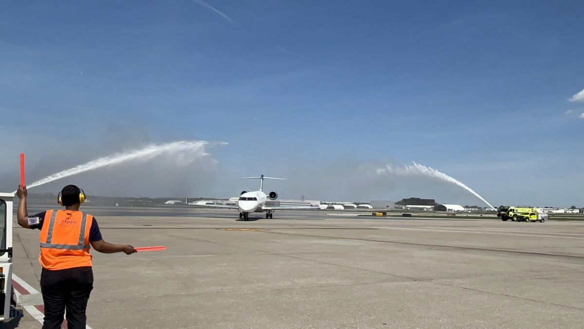 Photograph of a white regional jet taxiing on the runway that is flanked on both sides by airport fire trucks spraying high arcs of water over the aircraft. In the foreground, a ground crew member wearing a bright orange vest, earmuffs, and holding orange marshalling wands directs the aircraft toward the terminal.