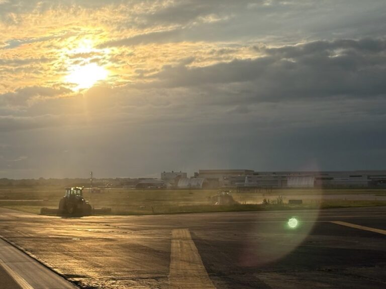 Airport maintenance vehicles mowing grass along the runway at sunrise, with golden sunlight filtering through low clouds and casting a glow on the tarmac.