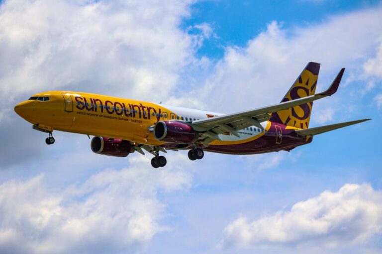 Sun Country Airlines Boeing 737 with maroon and gold livery approaches for landing against a backdrop of blue sky and scattered clouds. The aircraft features the Sun Country logo and 'Proud Sponsor of Minnesota Gopher Athletics' on the fuselage