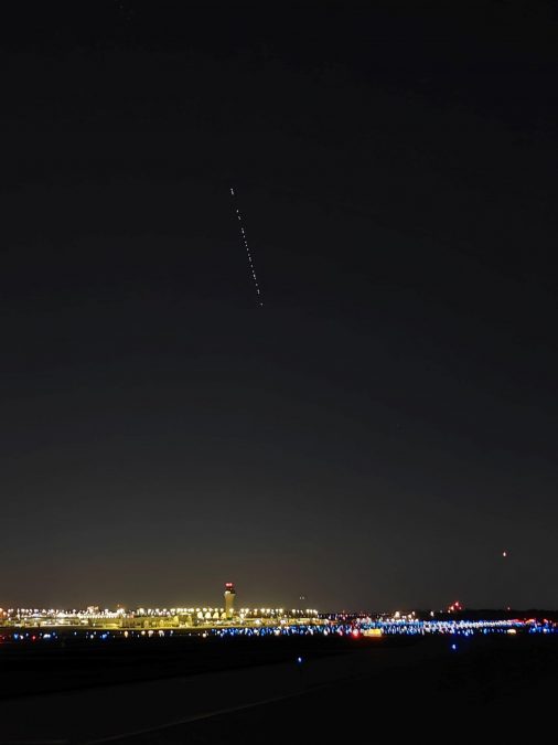 A nighttime view of an airport runway lit with blue and red lights, with a distant control tower illuminated. A visible string of Starlink satellites arcs across the dark sky in a dotted line formation.