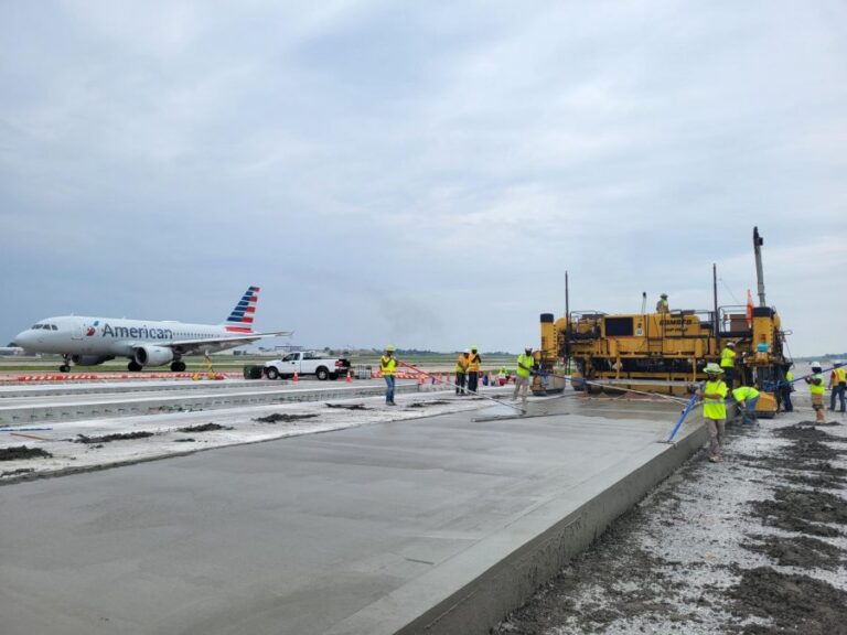 Airport runway construction in progress with a large yellow paving machine and workers in neon safety vests. An American Airlines jet is taxiing nearby in the background under a cloudy sky.