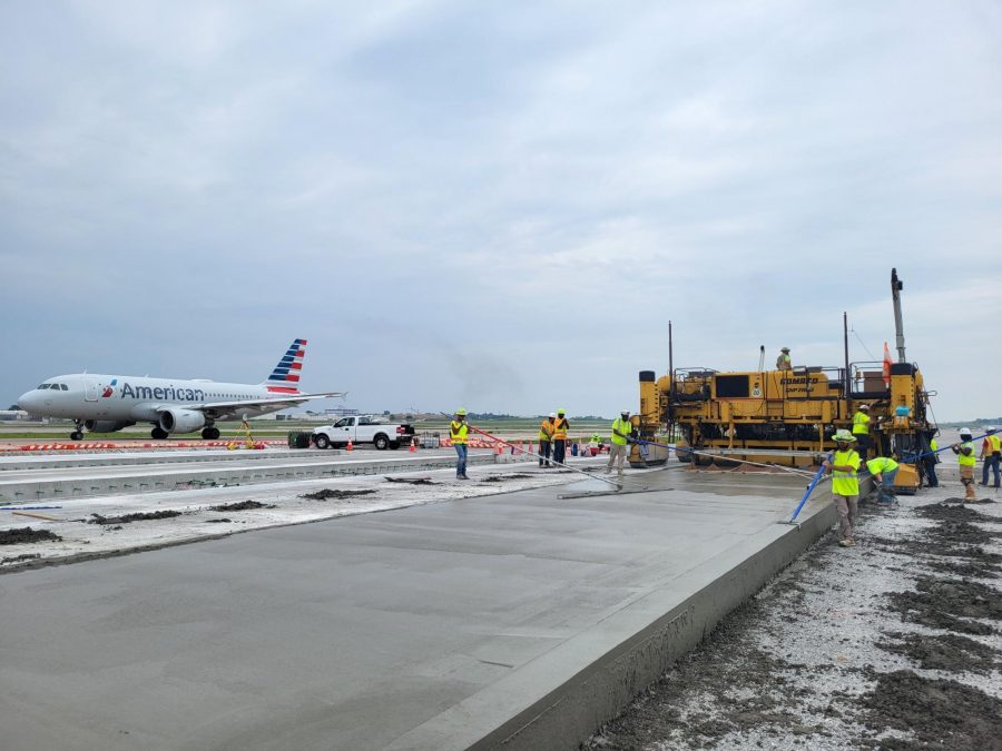 Airport runway construction in progress with a large yellow paving machine and workers in neon safety vests. An American Airlines jet is taxiing nearby in the background under a cloudy sky.