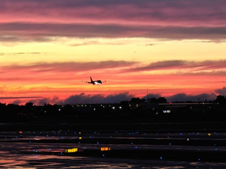 Commercial airplane approaching for landing against a vivid orange and purple sunset sky, with illuminated taxiway lights and wet runway reflections below.