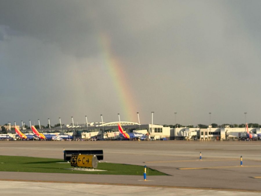 A faint rainbow appears in the gray sky above the airport terminal, where several Southwest Airlines planes are parked at their gates. The runway and taxiway signs are visible in the foreground.