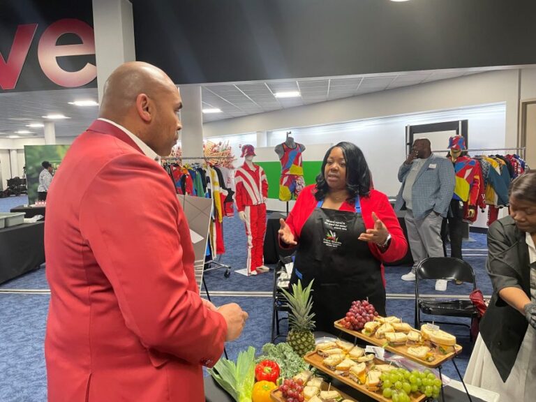 Vendor wearing a branded apron discusses catering offerings with an attendee at an airport event, with colorful apparel displays and sample food trays visible.