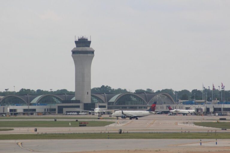 Daytime photograph of St. Louis Lambert International Airport’s main terminal, with its iconic curved roof design and prominent air traffic control tower rising in the center. The terminal’s arched windows reflect the mid-century modern architecture. Several Delta Air Lines aircraft are visible on the tarmac, parked at gates and taxiing on the runway. A row of flags flutters near the terminal entrance, and trees line the background under an overcast sky.