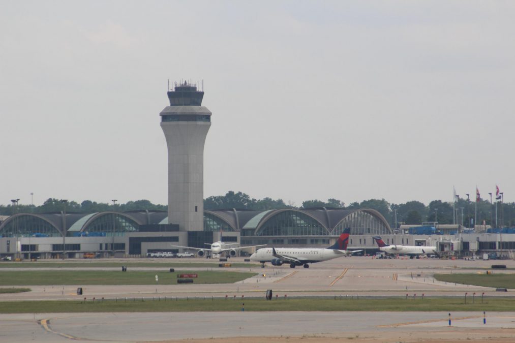 Daytime photograph of St. Louis Lambert International Airport’s main terminal, with its iconic curved roof design and prominent air traffic control tower rising in the center. The terminal’s arched windows reflect the mid-century modern architecture. Several Delta Air Lines aircraft are visible on the tarmac, parked at gates and taxiing on the runway. A row of flags flutters near the terminal entrance, and trees line the background under an overcast sky.
