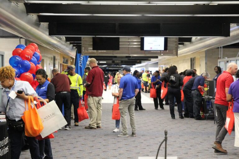 Airport terminal hallway filled with employees and vendors during an employee resource fair. People are talking at booths, carrying red tote bags, and walking past displays with balloons and informational materials.