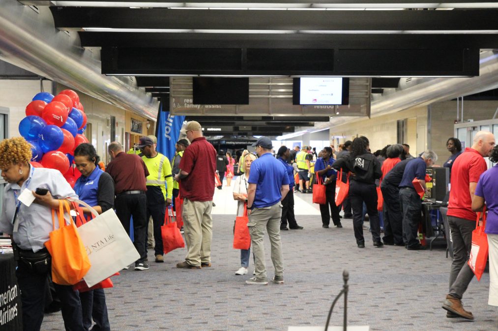 Airport terminal hallway filled with employees and vendors during an employee resource fair. People are talking at booths, carrying red tote bags, and walking past displays with balloons and informational materials.