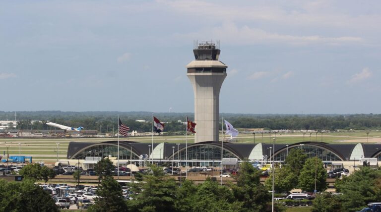 The STL airport control tower stands behind the terminal with curved roofs, surrounded by parked cars, flags, and trees, with an airplane taxiing on the runway in the background under a partly cloudy sky.