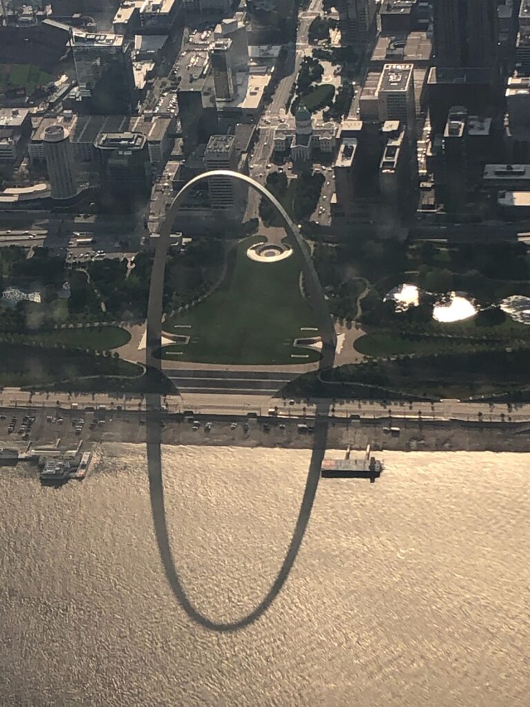 Aerial view of the Gateway Arch in St. Louis casting a large oval shadow over a green park and part of the city along the riverfront.