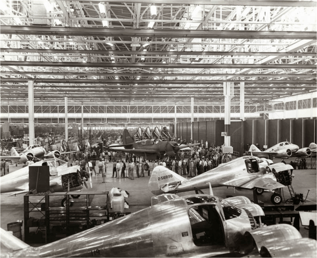 A large aircraft factory with numerous airplanes in various stages of assembly. Workers are gathered in groups around several planes under bright overhead lighting in the spacious industrial building.