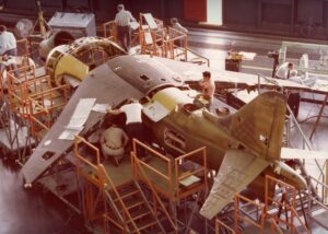 Engineers and technicians work on assembling and inspecting a partially constructed aircraft inside a large hangar, surrounded by scaffolding and tools.