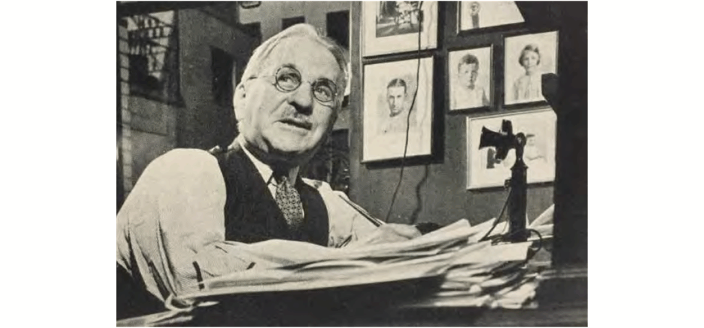 An older man wearing glasses and a vest sits at a cluttered desk, writing, with framed portraits hanging on the wall behind him and an old-fashioned telephone nearby.