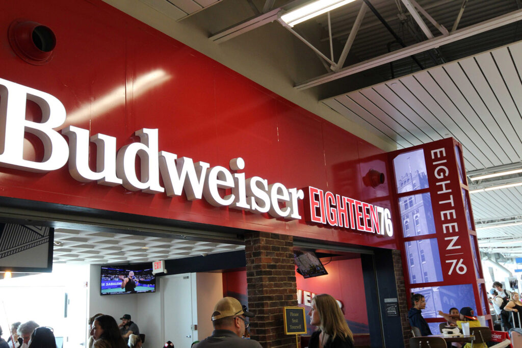 A Budweiser Eighteen76 sign at a concession stand in a stadium, with people ordering food and drinks below. The stand features red and white signage and exposed ceiling beams above.