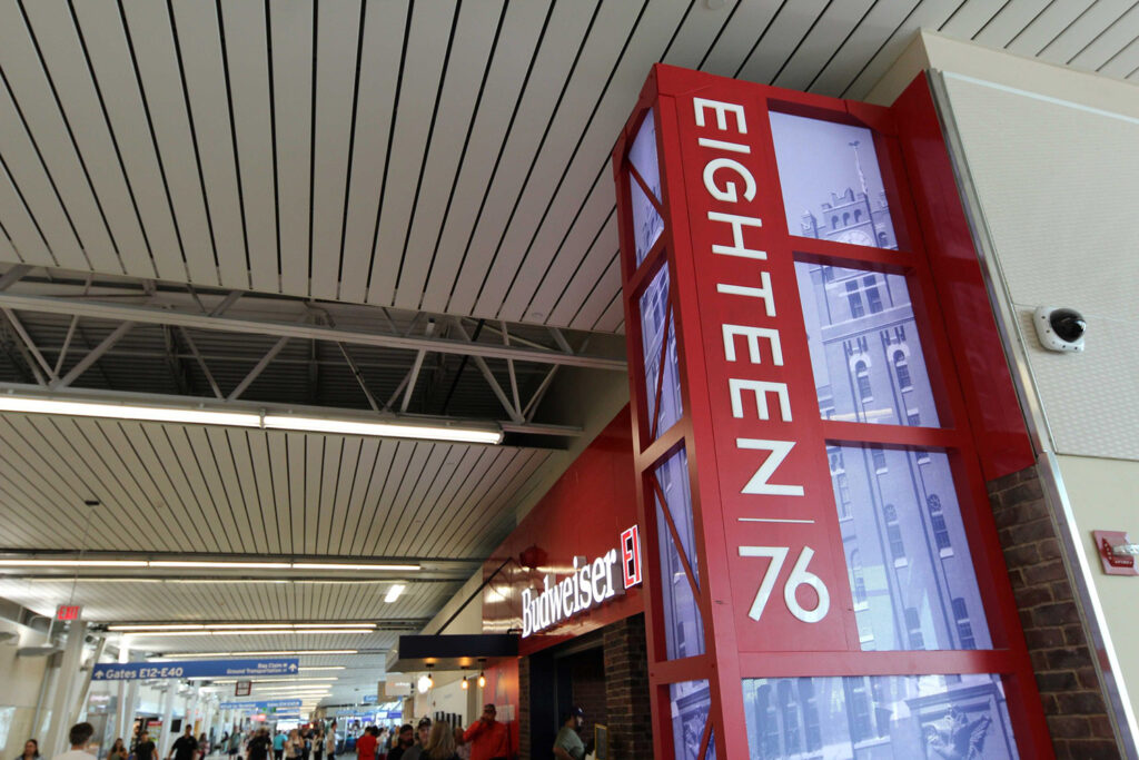A busy airport terminal with people walking, a red column displaying the numbers Eighteen 76, and a Budweiser sign above a bar or restaurant. Overhead, signs indicate gates and directions.