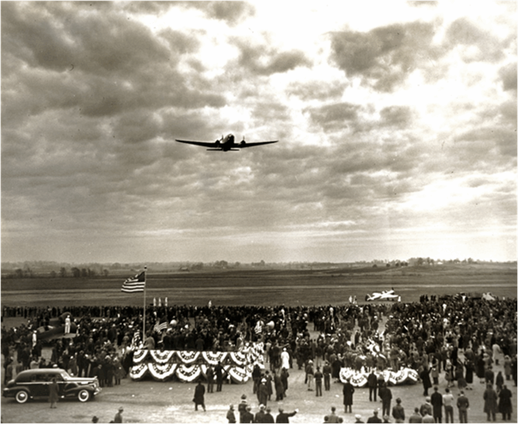 A large crowd gathers outdoors near decorated platforms and a US flag, watching an airplane fly low overhead under a cloudy sky. A vintage car is parked to the side.