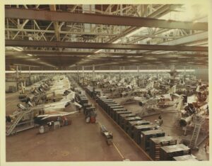 Large aircraft assembly line inside a spacious factory with rows of partially constructed fighter jets, maintenance platforms, machinery, workers, and various tools organized throughout the bright, high-ceilinged building.