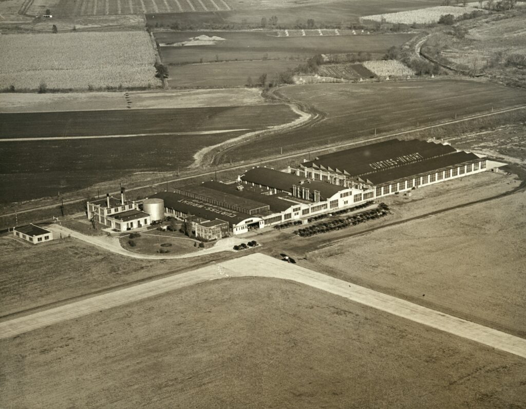 Aerial view of a large industrial building with multiple sections and a smokestack, surrounded by open fields, roads, and a few cars parked near the entrance. Farmland and trees are visible in the distance.