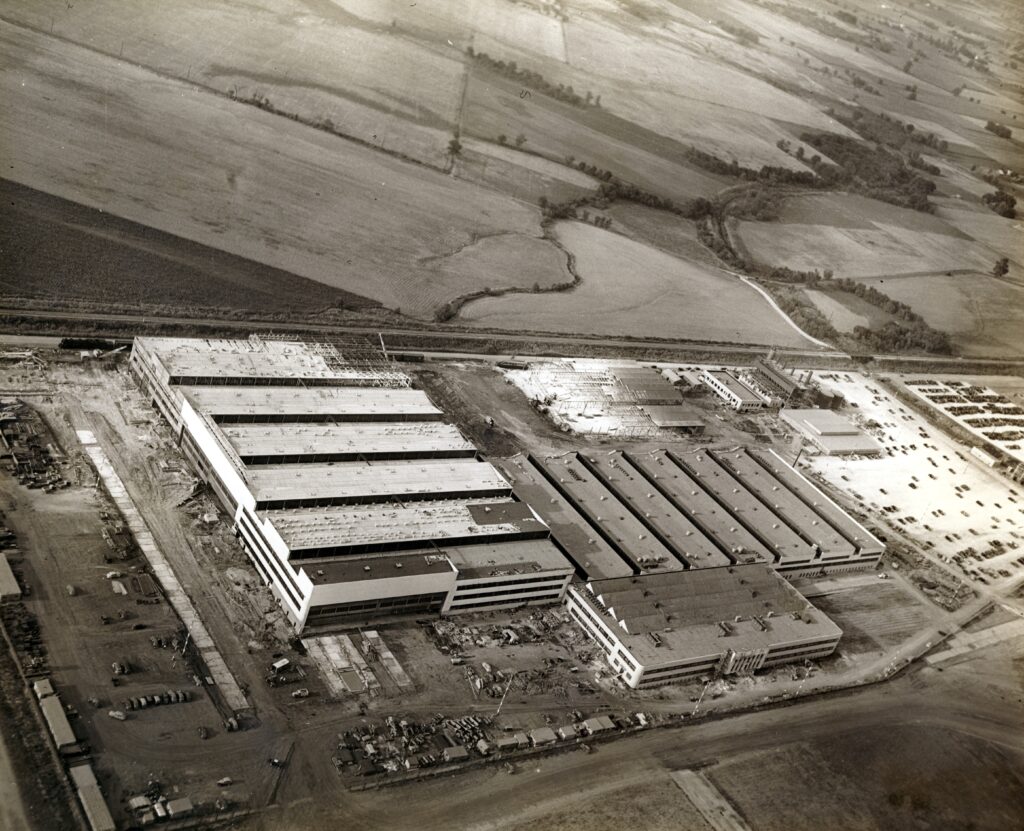 Aerial view of a large construction site with several partially completed industrial buildings surrounded by open fields and roads, with vehicles and construction equipment visible.