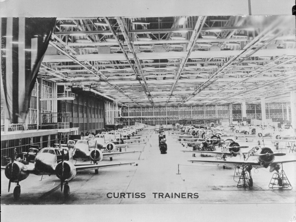 A large aircraft factory hangar filled with rows of partially assembled WWII-era Curtiss trainer airplanes. The words CURTISS TRAINERS appear at the bottom of the image.