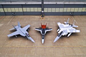 Three modern fighter jets are parked side by side on a concrete tarmac in front of a large hangar, viewed from above. The jets are different models and are painted in various colors.