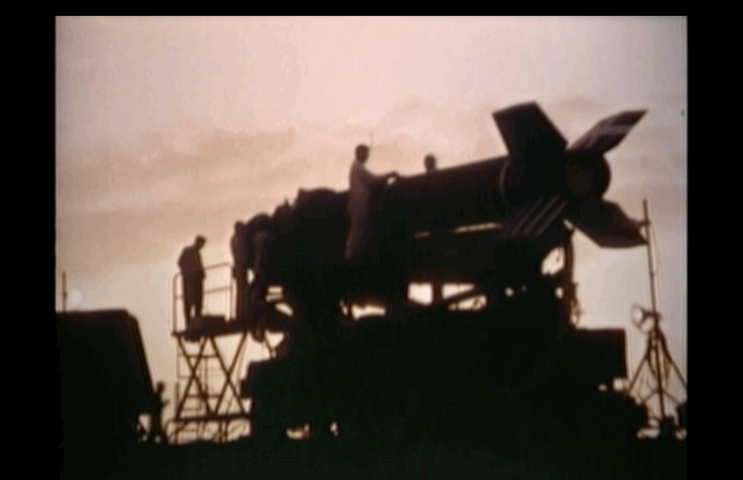 Silhouetted figures stand on scaffolding around a large rocket at sunset, preparing or inspecting it against a softly lit sky.