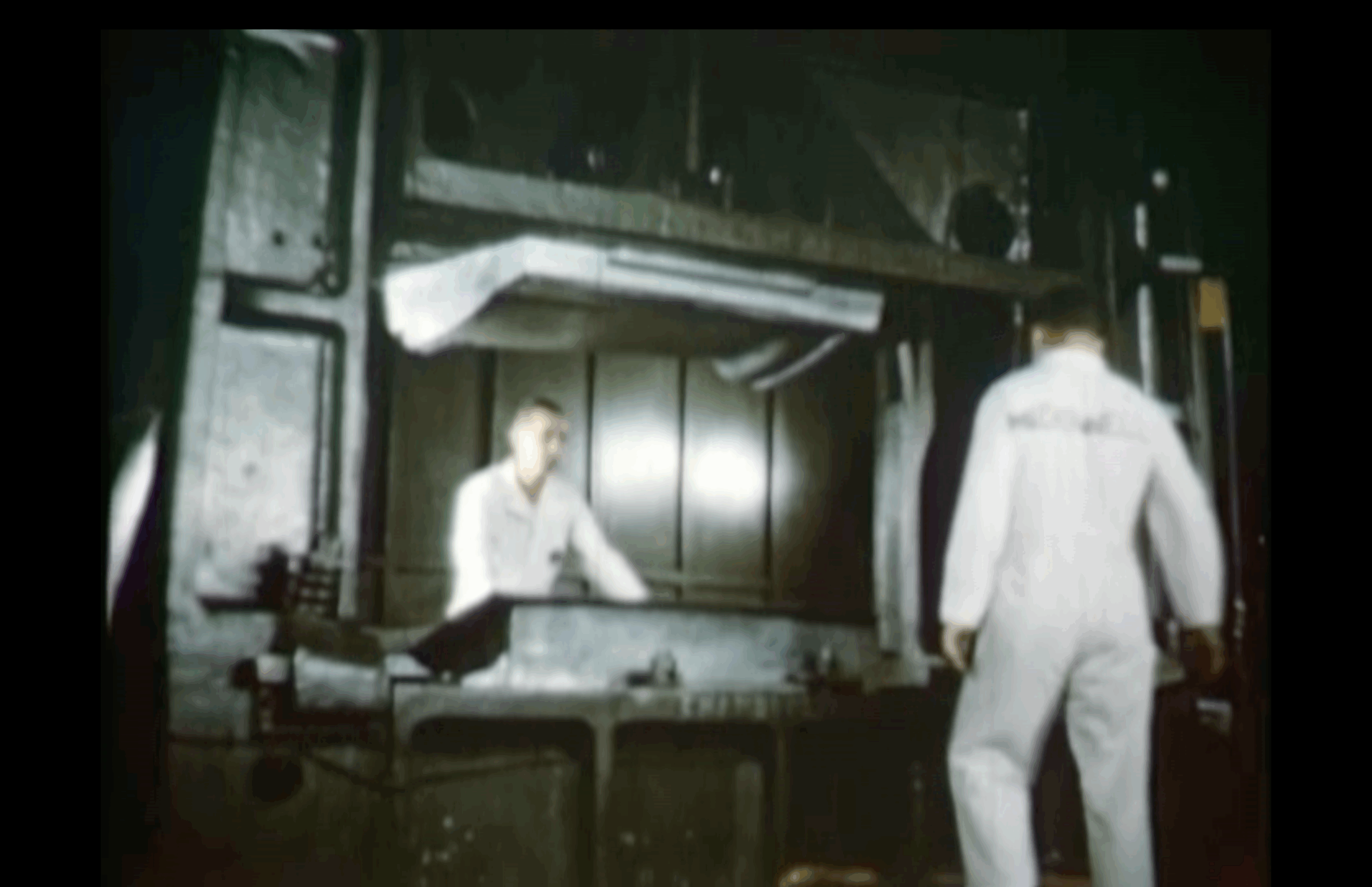 Two people in white lab coats stand in an industrial setting with large machinery and metallic surfaces in the background.