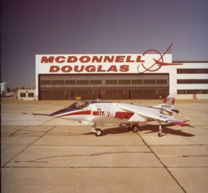 A white and red military jet labeled MARINES is parked on a tarmac in front of a large hangar with MCDONNELL DOUGLAS in bold red letters on the building.