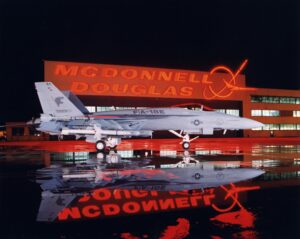 A U.S. Navy F/A-18E fighter jet is parked on a wet tarmac at night, reflecting on the ground, in front of a brightly lit McDonnell Douglas building with a glowing red logo and sign.