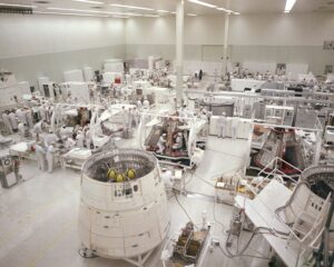A large, bright cleanroom with many engineers in white suits working on several spacecraft capsules and technical equipment, surrounded by tools and workstations.