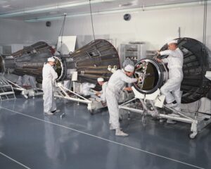 Several engineers in white cleanroom suits work on two large spacecraft capsules inside a bright, sterile laboratory. The capsules are mounted on wheeled stands, and various tools and equipment are visible around them.