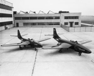 Two vintage U.S. Navy fighter jets are parked on a large concrete apron in front of a multi-story industrial building with many windows on an overcast day.