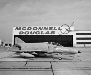 A McDonnell Douglas F-4 Phantom II jet is parked on a tarmac in front of a large hangar with MCDONNELL DOUGLAS written on it. The jet has visible markings and missiles attached under its wings.