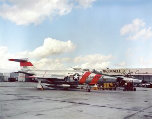 A silver U.S. Air Force jet with red markings is parked on a tarmac near a McDonnell Aircraft Corporation hangar under a partly cloudy sky. Ground crew and equipment are visible beside the aircraft.
