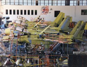 Several unfinished fighter jets in yellow primer are lined up in an aircraft assembly hangar, surrounded by scaffolding, tools, and workers. An American flag and sign reading “1000 for freedom” are displayed on the wall.
