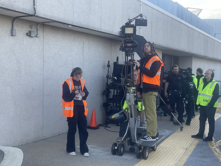 Film crew members in orange vests work outside a building. One person operates a camera on a dolly, while another checks their phone. Other crew and equipment are visible in the background.