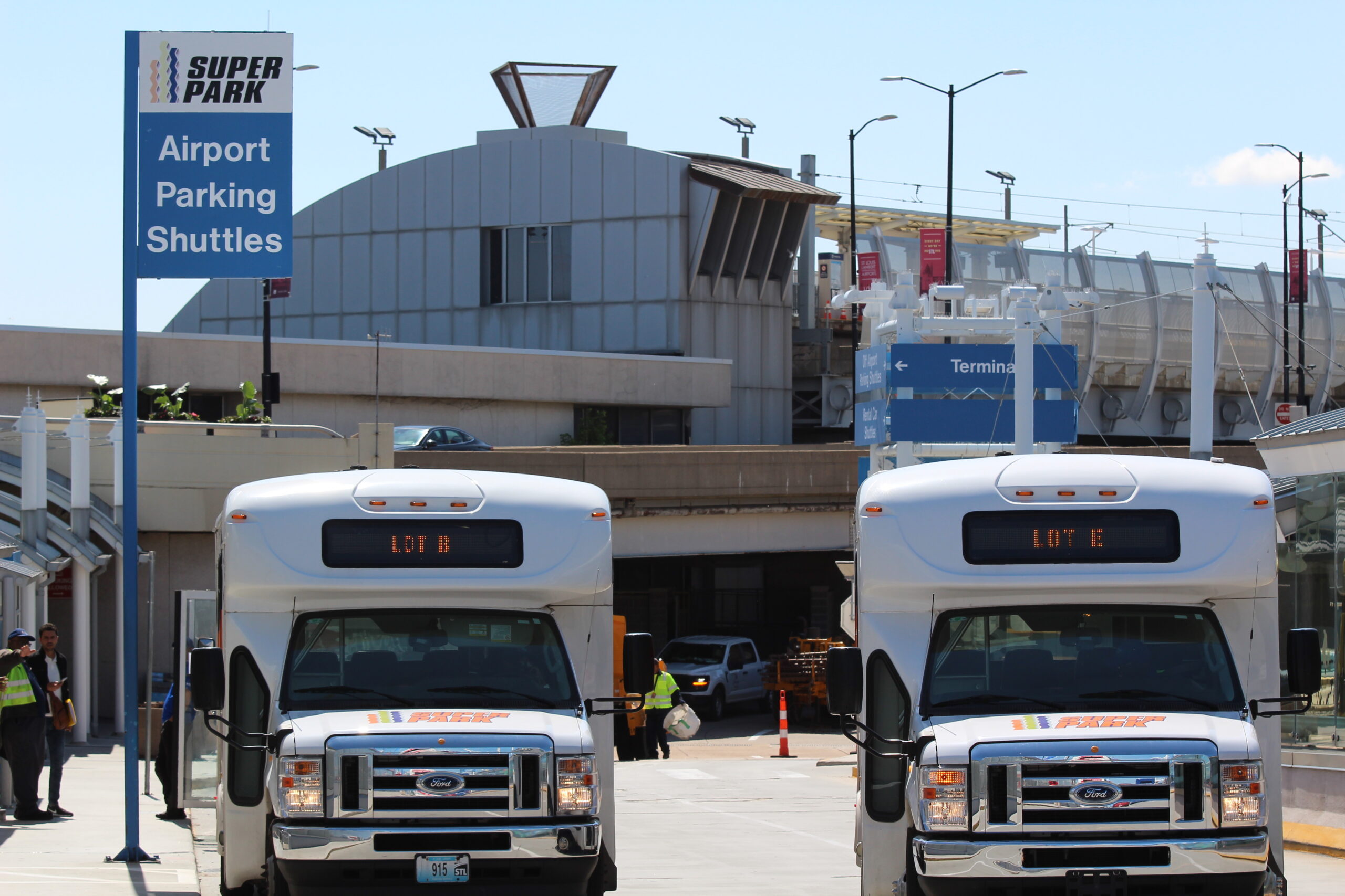 This is a phot of two Super Park parking shuttles, side-by-side, on Arrivals Drive at the Door 12 shuttle stop at Terminal One.