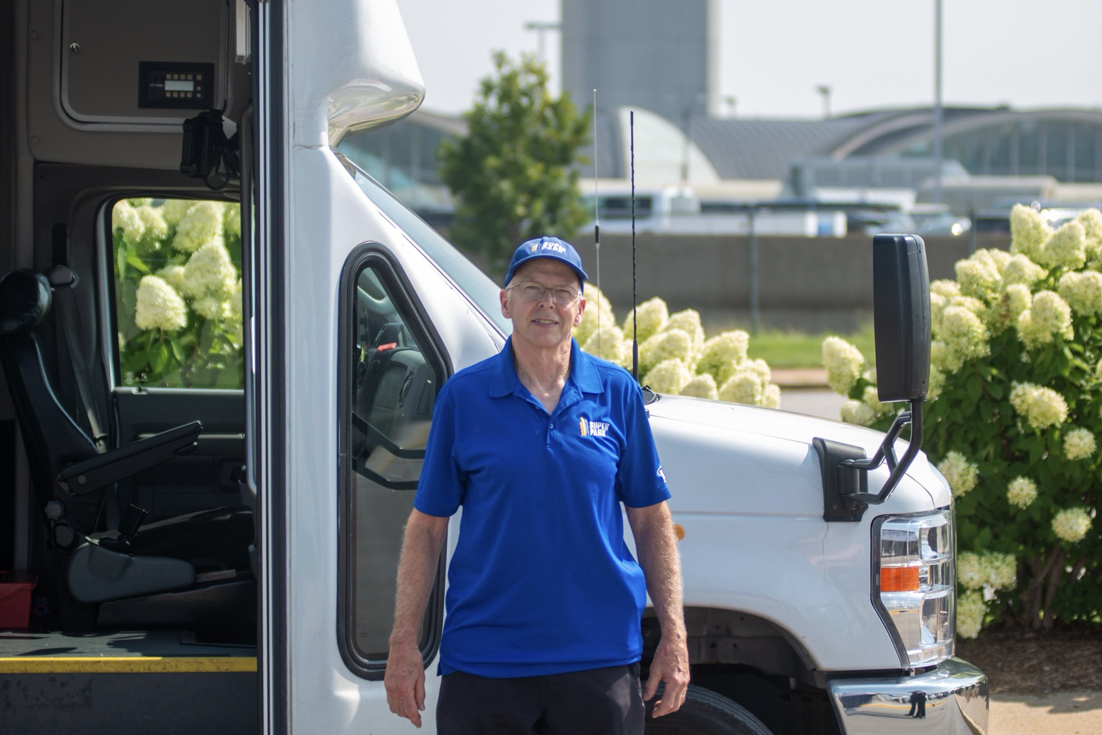 Michael Mayo a former member of the Super Park Facility Maintenance team stands in front of the cab of a Super Park shuttle bus at the airport's parking Lot A facility. In the background is the airport iconic Terminal 1 and the airport FAA control tower.