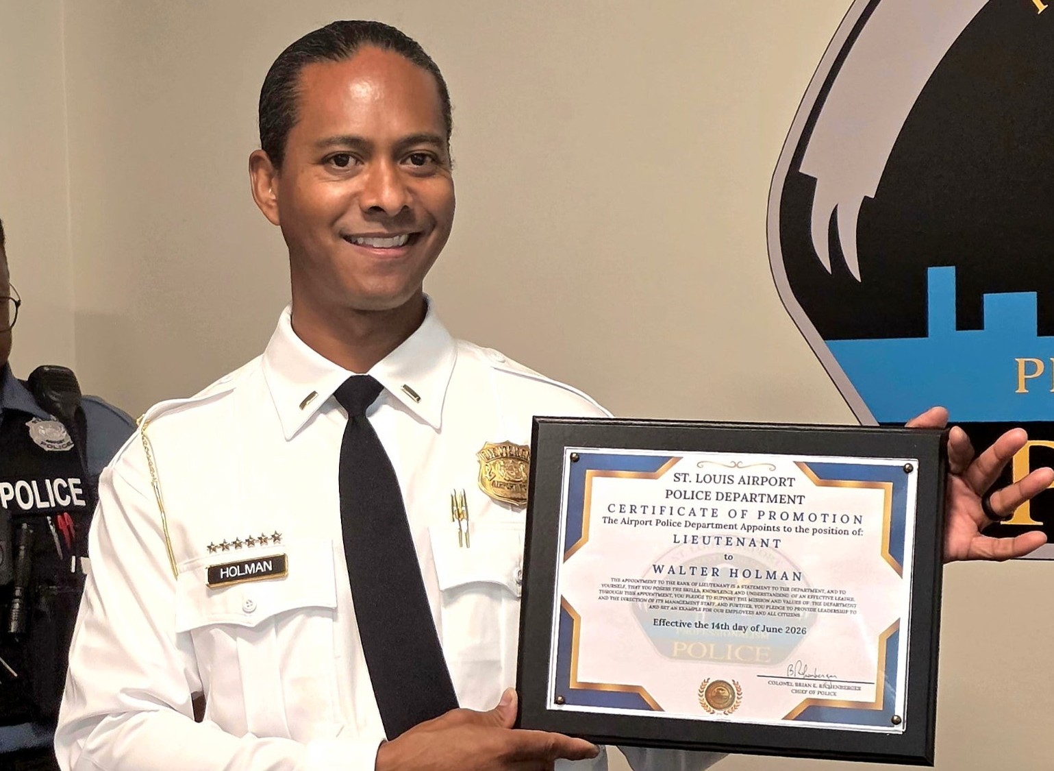 This photo shows newly promoted Lieutenant Walter Holman with his certificate of promotion. He is standing just to the left of the Airport Police Department's logo in the department's conference room.
