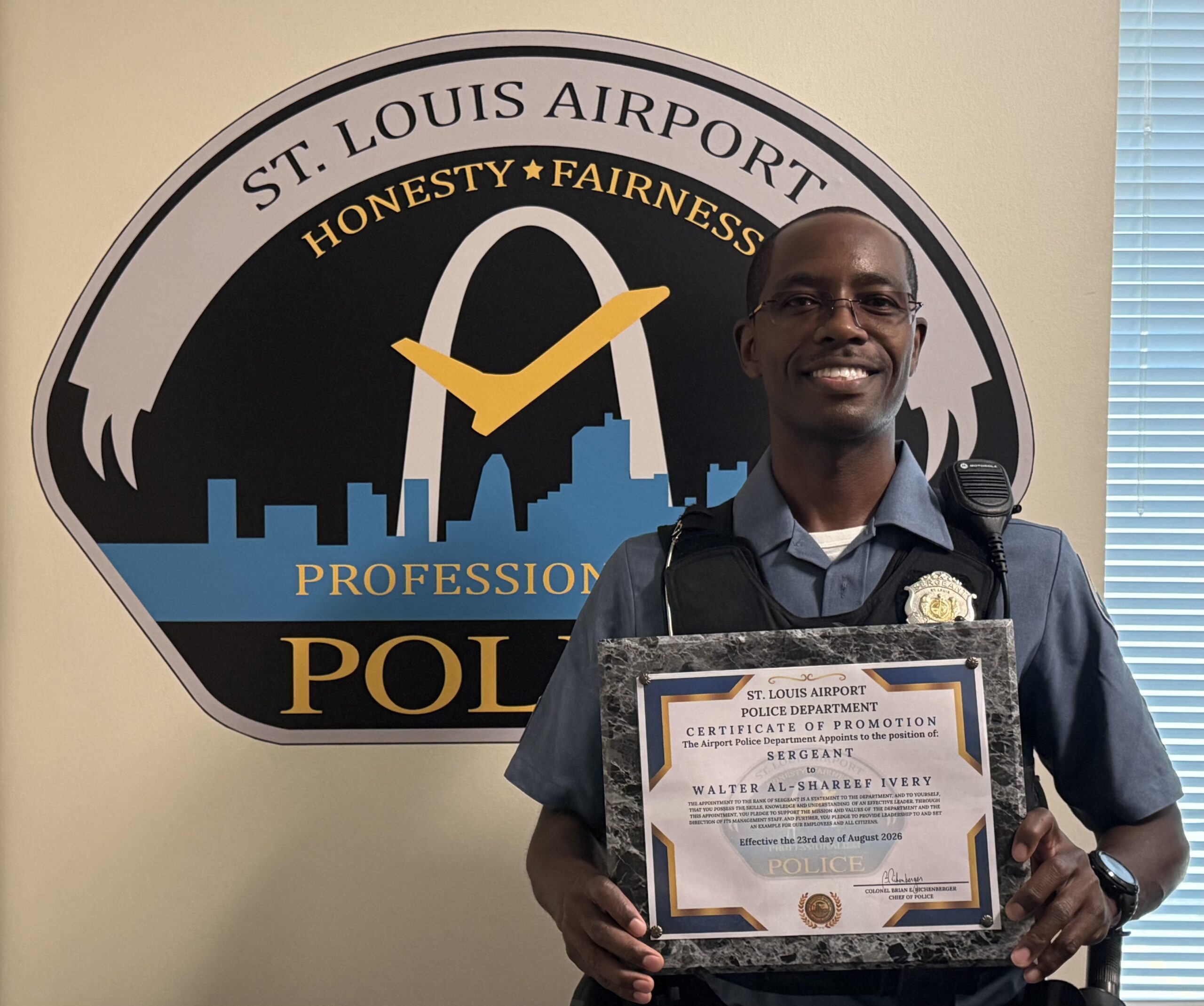 This photo is of newly promoted Sergeant Walter Ivery standing in front of St. Louis Lambert International Airport Police Department's wall logo in the department's conference room. He is standing with his promotion plaque.