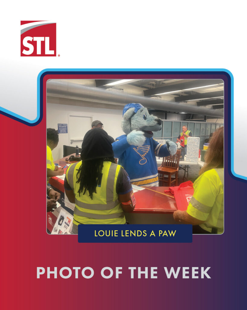 A person in a blue hockey mascot costume helps volunteers in safety vests at an event. Text reads, LOUIE LENDS A PAW and PHOTO OF THE WEEK, with the STL logo at the top left.