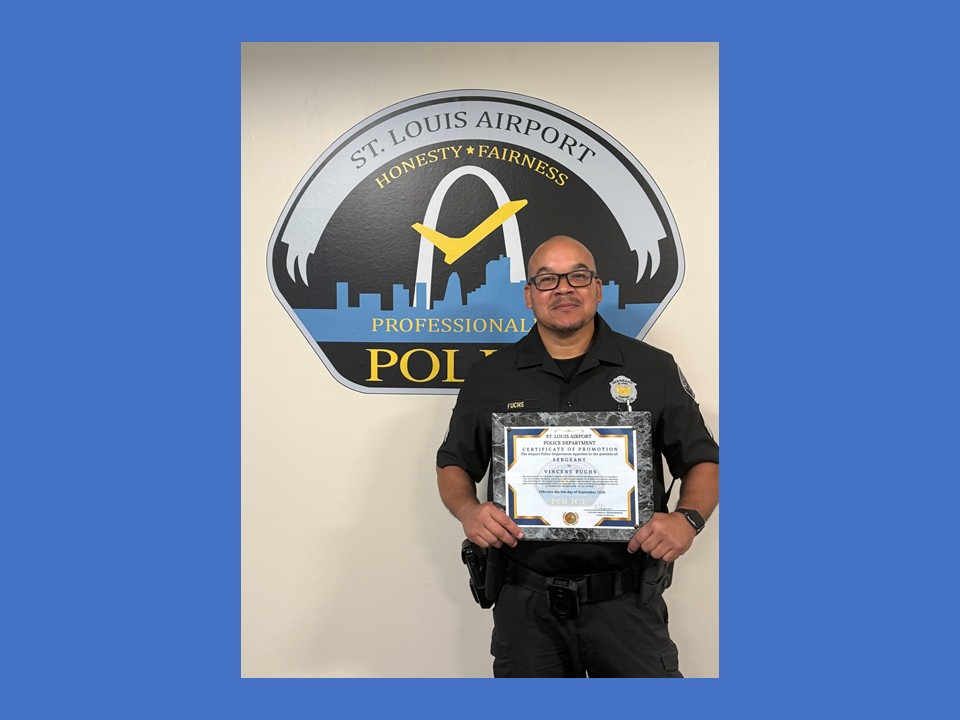 Airport Police Officer, Sgt. Vincent Fuchs standing in front of the STL Airport Police logo, which is mounted on the wall in the police department's conference room. He is holding his promotion certificate.