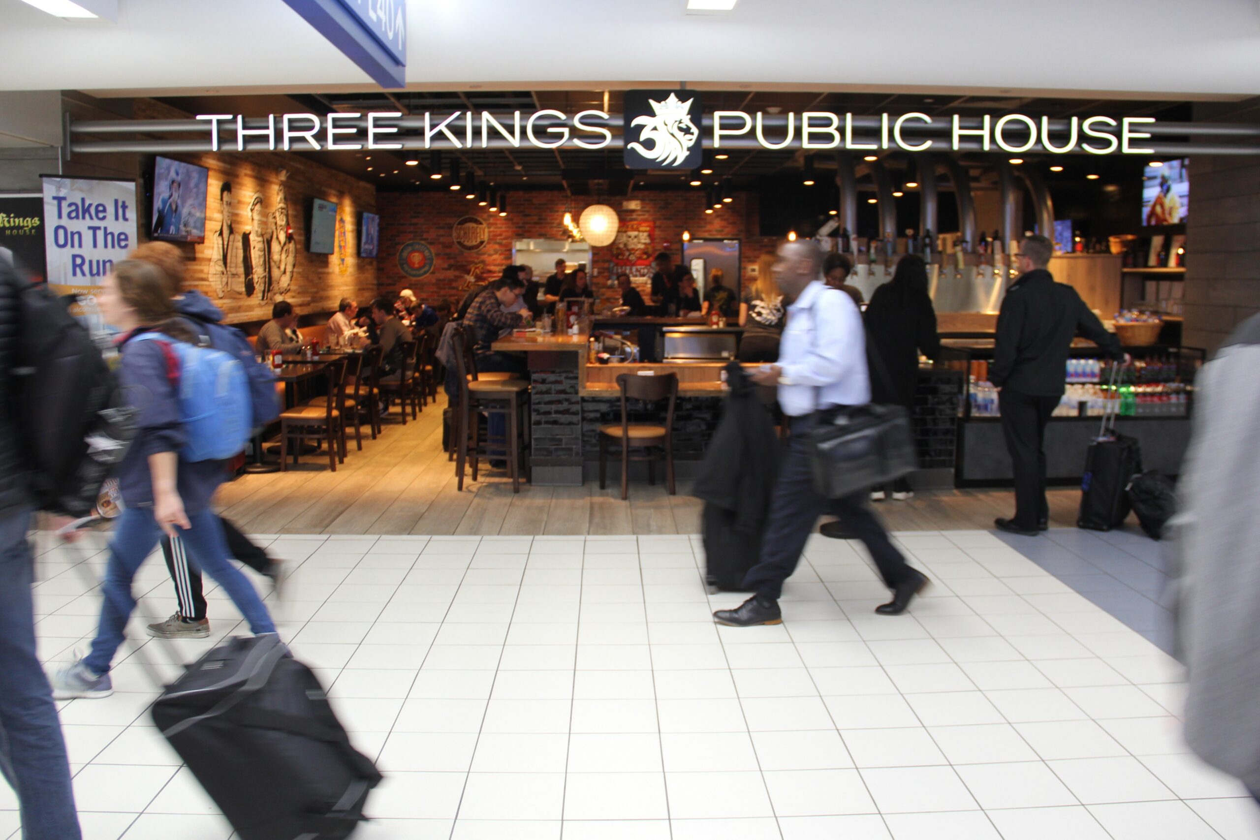 This is a photo of Three Kings Public House in STL's Terminal 2. It shows passengers walking past the location as others are seated in the restaurant eating .